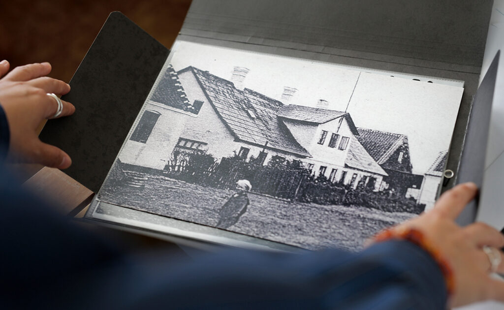 Hænder holder et album, der viser et gammelt sort-hvidt foto af et hus med stråtag og en person foran.